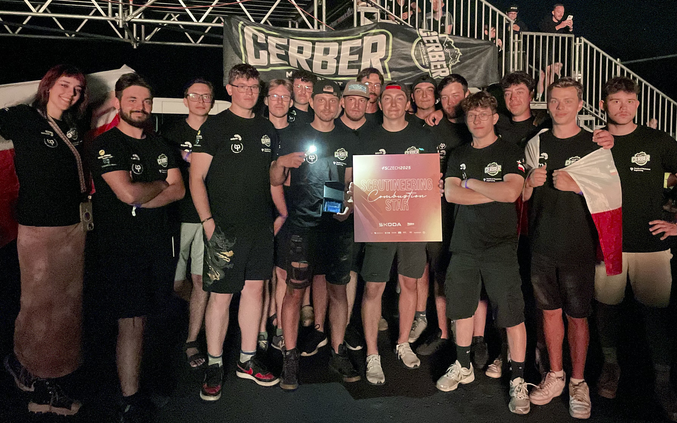 Grupa młodych osób pozujących na tle banera „CERBER Motorsport” nocą, dwie osoby trzymają tablicę z napisem „SCRUTINEERING Combustion START”, jedna osoba trzyma polską flagę.