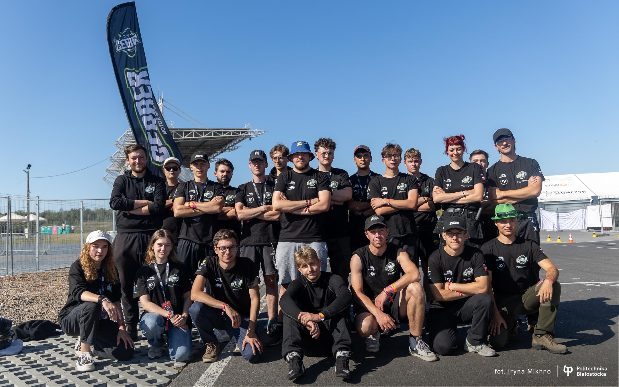 A group of about twenty young people pose together on a race track on a sunny day. They are all wearing black T-shirts with the Cerber Motorsport logo, some have caps or wristbands. In the background, there is a team flag, metal fencing, and parts of the track infrastructure.