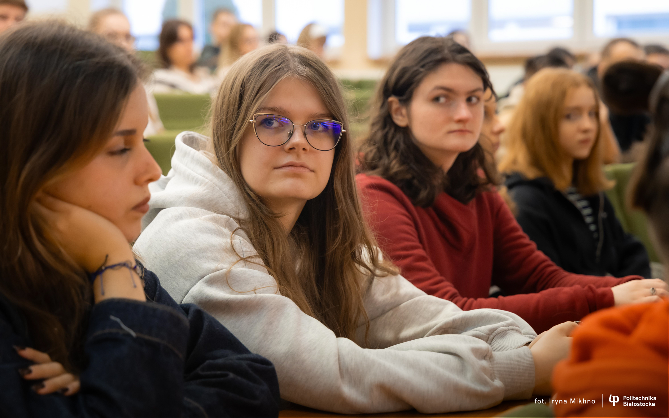 People sit in a lecture hall at tables with rows of chairs and bright windows in the background