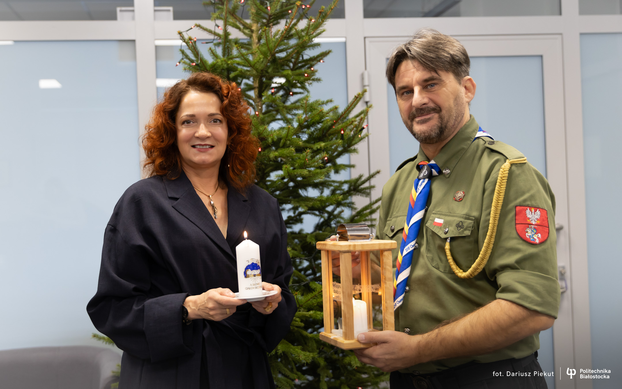 Two people stand by a Christmas tree holding a candle and a wooden lantern with a lit candle