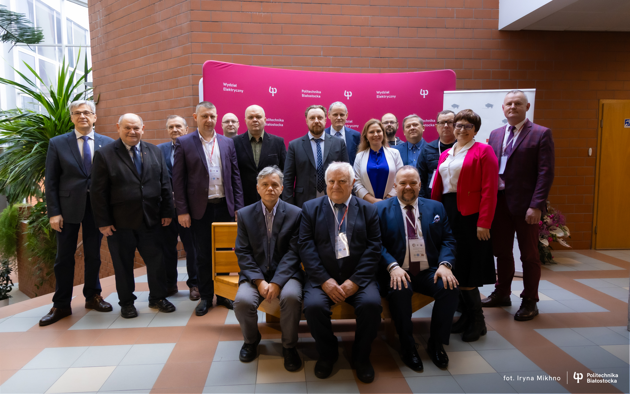 A group of people in formal attire posing for a group photo in front of a pink conference backdrop in a building lobby