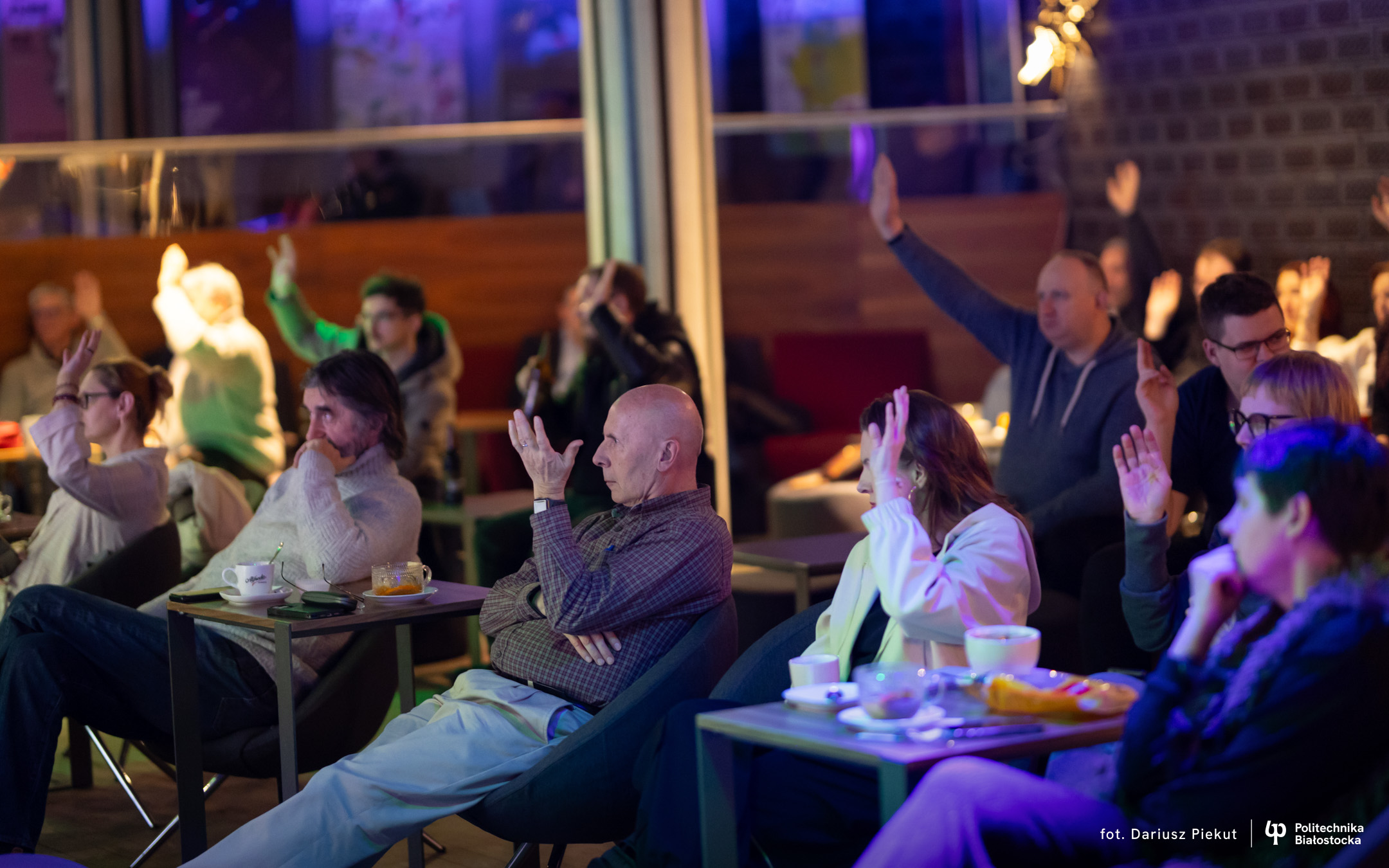 A group of people sitting at tables in a room lit with colourful lighting, raising their hands during a meeting or discussion event