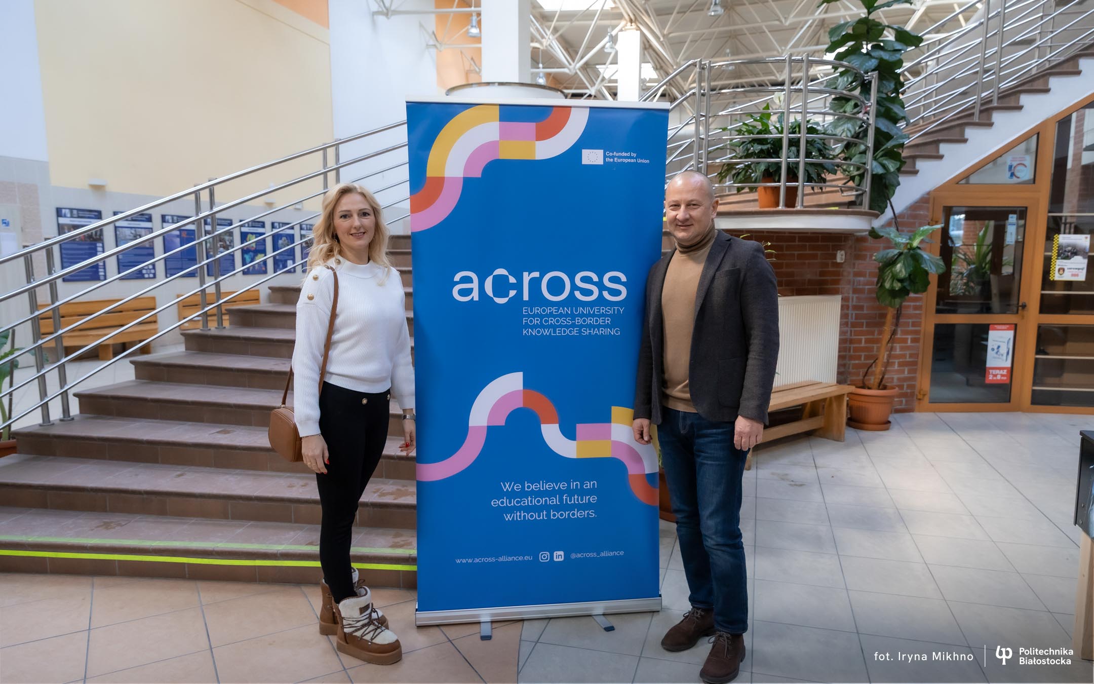 Two people standing next to a blue ACROSS project roll‑up banner in a building lobby with metal stairs and plants