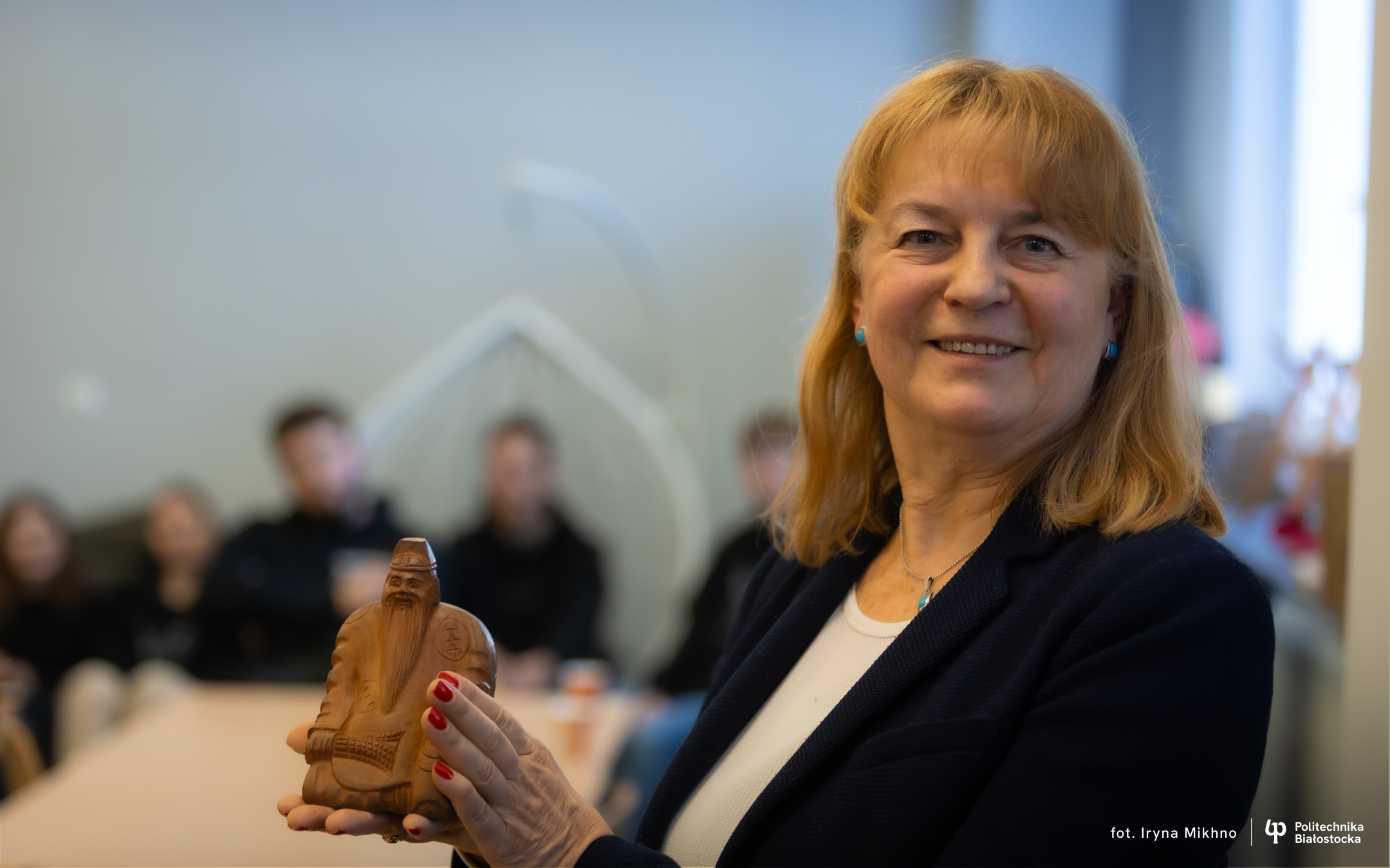 A person holding a small sculpture and presenting it to a group of participants seated at a table in a classroom setting